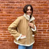 Campera de cuero ovino color tostado con interior de piel sintética blanca. Tiene cuello alto con solapas y cierre frontal con cremallera.