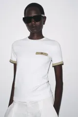 Camiseta blanca de corte slim fit, cuello redondo y manga corta. Detalle de acabados en tricot con hilo metalizado color dorado en mangas y bolsillo.