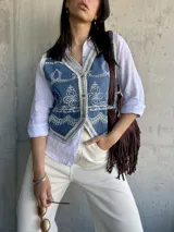 Chaleco de jean azul con bordados florales blancos y ribete de encaje. Cierre frontal con botones.