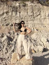 Pantalón largo color beige claro, de tejido ligero con textura, tiro alto y cintura elástica. Presenta detalles deshilachados o con terminación cruda en los bordes verticales de la pierna, creando un efecto drapeado y fluido.