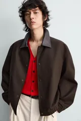 Cazadora de algodón con estructura canvas, color marrón oscuro, de corte relajado. Presenta cuello solapa de cuero negro, cierre frontal con botones a presión, bolsillos de vivo en cadera y puños y bajo elásticos.