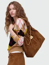 Bolso tipo hobo o bucket de gamuza marrón, con frunce en la parte superior y detalles de costura en contraste. Cuenta con una cadena dorada decorativa y un dije metálico con logo.