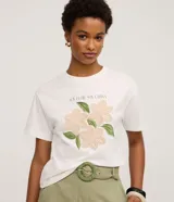 Remera blanca de algodón, con cuello redondo y manga corta. Presenta un estampado frontal con la frase "SOY FLOR. SOY LATINA" y un diseño floral con flores en relieve y hojas bordadas.