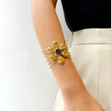 Pulsera artesanal de bronce con diseño de flor central compuesta por piedras blancas de forma ovalada. Presenta una estructura de múltiples hilos metálicos que forman un brazalete tipo cuff.