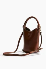 Cartera tipo balde color marrón con asa de mano rígida y correa bandolera ajustable y extraíble. Cierre con trabilla y adorno metálico dorado.