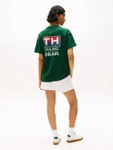 Remera verde de algodón con cuello redondo y corte amplio. Presenta el logo de Tommy Hilfiger en el pecho y un logo oversize en la parte trasera. Tiene mangas cortas con hombros caídos y un parche con logo en la manga.