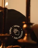 Remera negra de algodón con estampa circular en el frente que incluye un corazón con llamas, la palabra 'LUXO' y la frase 'TAROT x LOS AMANTES'.