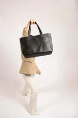 Cartera tipo tote de cuero graneado color negro, con textura uniforme y suave al tacto. Tiene manijas de 55 cm, bolsillo frontal con broche imantado, bolsillo trasero con cierre metálico, cierre principal metálico, forro interior y tres bolsillos internos (uno con cierre, uno plaqué y uno tipo sobre con solapa y botón).