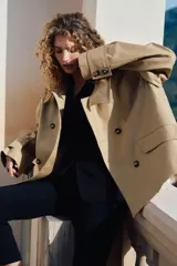 Gabardina corta color beige con doble botonadura, cuello clásico, charreteras en los hombros, bolsillos delanteros con solapa, mangas anchas con trabillas ajustables y pliegue en la espalda.
