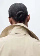 Abrigo tipo trench color beige, largo, con cuello de solapa, trabillas en los hombros y mangas largas abullonadas con trabillas en los puños. Incluye cinturón desmontable y bolsillos laterales.