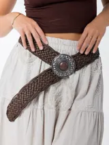 Cinto marrón trenzado con hebilla circular plateada con piedra marrón al centro.