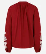 Blusa color borgoña de batista con cuello en V y botones. Presenta bordados florales blancos en el frente y las mangas largas con puños elásticos.
