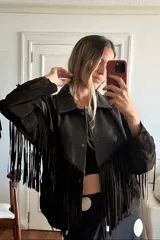 Chaqueta tipo bomber de cuero ovino marrón desgastado y gamuza color chocolate, con flecos largos en el canesú frontal y trasero. Presenta cuello solapa y cierre frontal con broches forrados.