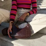 Cartera de hombro color marrón oscuro con textura de cocodrilo (efecto croco) y acabado brillante. Tiene forma de media luna y asa corta.