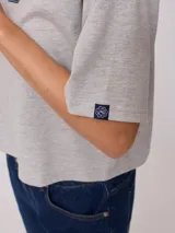 Remera gris de algodón con corte cropped y calce holgado. Tiene cuello redondo y mangas cortas con costuras reforzadas en color coral. Presenta un pequeño logo de la marca Rusty en el pecho.