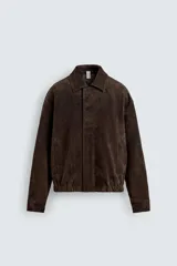 Cazadora de hombre de corte relajado confeccionada en piel con acabado ante, color marrón oscuro. Presenta cuello solapa, manga larga con puños abotonados a presión, bolsillos de vivo en cadera, bolsillo interior y bajo con elástico. Cierre frontal oculto por solapa con botones a presión.