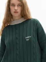 Buzo verde oscuro de tejido de punto con cuello alto y mangas largas. Tiene un pequeño logo bordado en el pecho y puños y cuello en color beige con rayas horizontales.