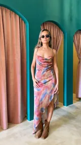 Vestido midi con estampado tie-dye en tonos rosados, azules y beige. Tiene escote drapeado, breteles finos y falda irregular con abertura lateral.