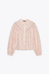 Camisa rosa pálido con bordados perforados, cuello con volante y mangas largas abullonadas. Cierre frontal con botones a tono.