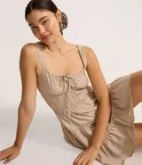 Vestido corto color beige, con escote recto, breteles medianos y lazo decorativo en el centro del busto. Presenta costura fruncida en la cintura y falda con babados en capas.