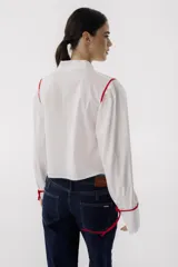 Camisa blanca de corte cropped con cuello mao y mangas largas abullonadas con lazos rojos en los puños. Presenta detalles de ribetes rojos en los hombros y lazos rojos en el cuello.