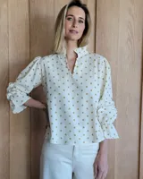 Blusa color beige con cuello con volados, mangas largas abullonadas con puños elásticos y cierre con botones en la parte delantera.