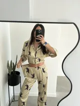 Pijama de dos piezas color beige con estampado de tigres negros y ribetes negros. Incluye camisa de manga larga con cuello y botones, y pantalón largo de corte recto.