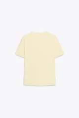 Camiseta marrón de algodón de manga corta y cuello redondo. Presenta un bolsillo en el pecho con un broche metálico dorado con diseño intrincado.
