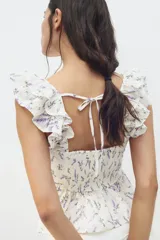 Blusa blanca con estampado floral en tonos lavanda, de tela ligera, con escote recto, breteles anchos con volados, espalda escotada con tiras finas horizontales que se atan en la parte superior, sección de punto smock en la espalda baja, costura fruncida en la cintura y peplum con vuelo.