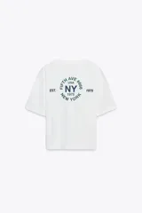 Camiseta blanca de algodón con cuello redondo, corte oversize y mangas cortas. Estampado frontal con la frase "New York" en azul.