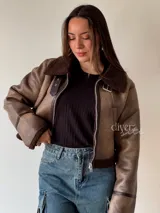 Campera corta color marrón con cuello, puños y ruedo de piel sintética. Cierre frontal con cremallera y detalles de hebillas en los laterales.