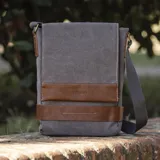 Morral gris de tela canvas con detalles en cuero marrón. Tiene correa larga regulable, tapa con broches magnéticos, bolsillo tipo guantera bajo la tapa, división interna con velcro y bolsillo trasero con broche magnético.