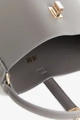 Cartera gris tipo bucket con asa corta y correa larga extraíble. Tiene cierre con trabilla y herraje metálico dorado. Interior forrado.