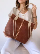 Bolso tipo tote de cuero marrón con costuras blancas a la vista y tiras decorativas. Asas de cuero color crema con detalle de trenzado. Forrada en tela.