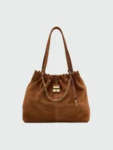 Bolso tipo hobo o bucket de gamuza marrón, con frunce en la parte superior y detalles de costura en contraste. Cuenta con una cadena dorada decorativa y un dije metálico con logo.
