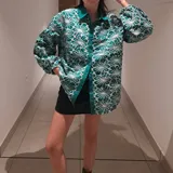 Camisa verde con estampado de flores blancas, cuello clásico, cierre con botones y mangas largas abullonadas con puños elásticos.