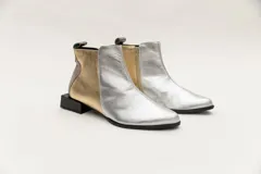 Botín corto de cuero color bronce con detalle de pieza de cuero charolada negra en el talón, cierre lateral y taco geométrico.