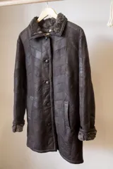 Abrigo de cuero marrón con cuello de piel sintética, cierre con botones y bolsillos laterales. Diseño con paneles geométricos y costuras a la vista.