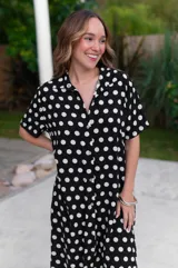 Camisola larga de muselina negra con estampado de lunares blancos, cuello camisero y manga corta. Presenta un corte holgado y abotonadura frontal completa.