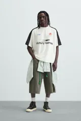 Camiseta blanca de corte holgado con mangas cortas negras. Presenta un logo azul en el pecho izquierdo, un escudo rojo con un caballo en el pecho derecho y la inscripción "Victory Games" en negro en el centro.