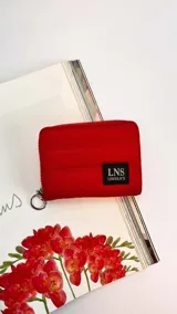 Billetera corta roja con cierre alrededor y tapa. Presenta un diseño capitoneado y un parche negro con el logo "LNS LINCOLN'S". Incluye una correa de mano desmontable.