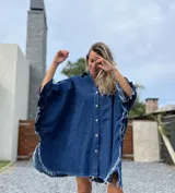 Camisola tipo poncho en jean azul oscuro con cuello camisero, cierre frontal con botones, mangas cortas amplias con volados y ruedo con flecos.