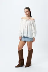 Blusa blanca de algodón con escote off-shoulder, mangas largas con puños elásticos y detalles de bordado y puntillas en escote y mangas.