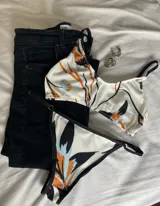 Conjunto de bikini blanco con estampado de hojas en tonos naranja, negro y celeste. El corpiño es de corte triángulo con tirantes finos y la bombacha es tipo tanga con laterales finos.