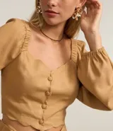 Blusa cropped color beige con escote corazón, mangas largas abullonadas con puños elásticos, botones frontales y detalle de volados en el ruedo.