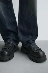 Mocasín negro con flecos. Parte superior lisa con efecto desgastado. Detalle de antifaz y flecos con puntas metálicas en el empeine. Horma cuadrada. Suela con volumen.