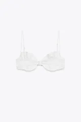 Top de bikini blanco con aros y tirantes finos ajustables. Tiene volantes en el pecho y un lazo negro en el centro. Cierre trasero con gancho metálico.