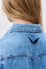 Camisa de denim celeste con cuello clásico, cierre frontal con botones a presión, dos bolsillos con solapa en el pecho y mangas largas con puños abotonados.