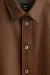 Camisa de corte oversized en denim de algodón rígido revestido, color marrón oscuro. Presenta cuello inglés, tapeta clásica con botones de metal y canesú en la espalda. Tiene hombros caídos, mangas largas con puños abotonados y bajo redondeado.