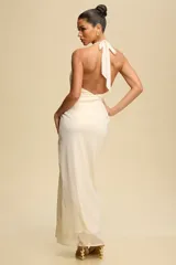 Vestido largo de fiesta color crema, con escote halter y espalda descubierta. Presenta un diseño drapeado con abertura en la falda y detalle de aro dorado en el cuello.